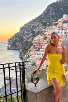 a woman in a yellow dress standing on a balcony next to the ocean and buildings