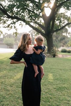 a woman in a black dress holding a toddler on her shoulders and looking off into the distance