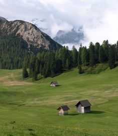 two small houses in the middle of a green field with mountains in the background and clouds