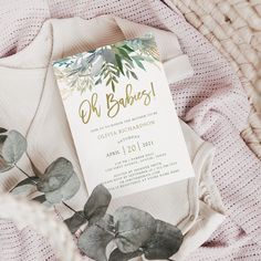 a white card with greenery and gold foil is on top of a pink blanket