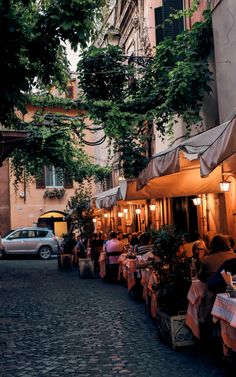 people sitting at tables in an alleyway with cars parked on the side walk behind them