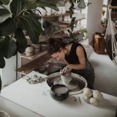 a woman sitting at a table making something out of clay on a potter's wheel