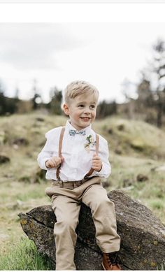 a little boy sitting on top of a rock wearing suspenders and a bow tie