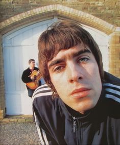 a young man with an acoustic guitar in front of a garage door and another person behind him