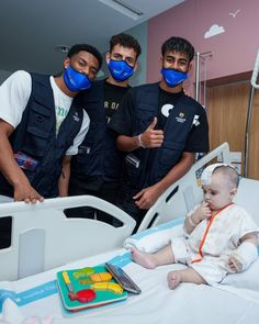 three men and a baby in a hospital bed wearing face masks while giving the thumbs up