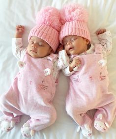 two baby babies laying next to each other on a white bed wearing pink outfits and hats