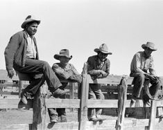 four men sitting on top of a wooden fence in cowboy hats and jeans, all looking at the camera