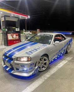 a silver car with blue stripes parked in front of a gas pump at night time