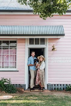 a man and woman standing in front of a pink house with the door open to their home