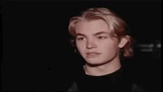 a man with blonde hair wearing a black shirt and looking at the camera while standing in front of a dark background