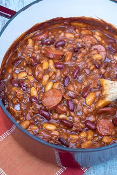 a pot filled with beans and sausages on top of a red table cloth next to a wooden spoon