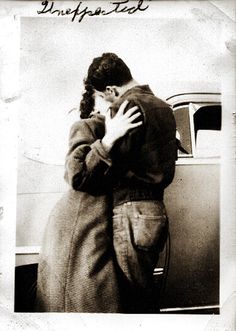 an old black and white photo of a man kissing a woman in front of a car