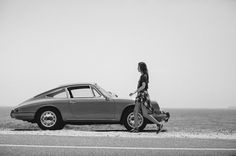 a woman standing next to a parked car on the side of the road near the ocean