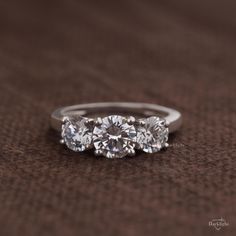 three stone diamond ring sitting on top of a brown clothed surface with the middle one in white gold and the other in silver