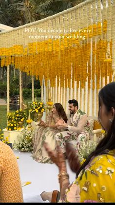 two women sitting on a couch in front of a man and woman with flowers hanging from the ceiling