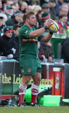 a rugby player holding a ball in his right hand while standing on the field with people watching