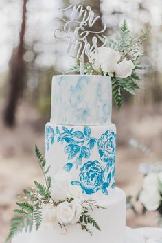 a blue and white wedding cake with flowers on top is sitting on a table in the woods