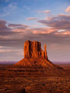 a large rock formation in the middle of a desert with mountains and clouds behind it