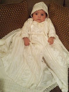 a baby wearing a white dress and bonnet is sitting on a couch with a blanket