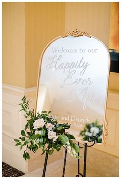 a welcome sign with flowers and greenery in front of it on a stand next to a wall