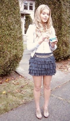 a woman standing in front of some bushes holding a cell phone and wearing a skirt