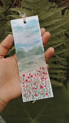 a hand holding a piece of paper with watercolor flowers on it and fern leaves in the background