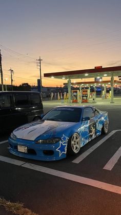 a blue sports car parked in front of a gas station at night with the sun setting