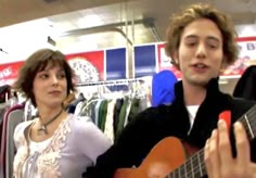 a man playing an acoustic guitar next to a woman in a clothing store while she looks at the camera