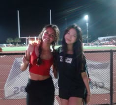 two women standing next to each other in front of a tennis court at night time