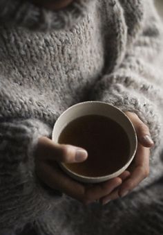 a woman holding a cup of coffee in her hands while wearing a gray knitted sweater