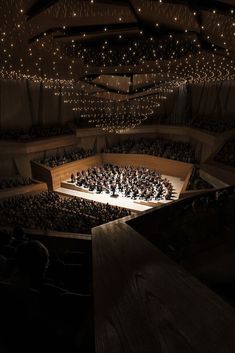 an auditorium filled with lots of people sitting on top of a wooden stage and lights hanging from the ceiling