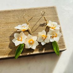 white and yellow flowers are hanging from gold earwires on a wooden board with green leaves