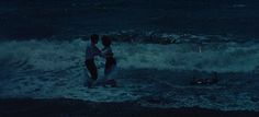 two people standing in the ocean at night with their arms around each other and one person on a surfboard