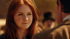 a close up of a person with red hair and a man in the background looking at her