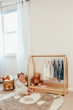 a baby sitting on the floor in front of a wooden shelf with clothes hanging from it