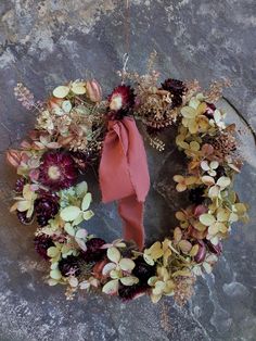 a wreath with dried flowers and a pink ribbon hanging on a rock wall, surrounded by foliage