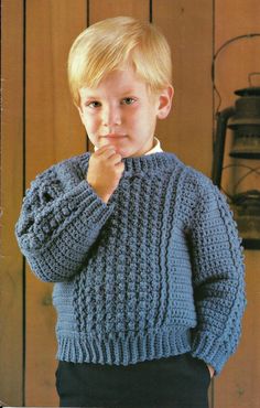 a young boy wearing a blue sweater and holding his hand to his mouth while standing in front of a wooden wall