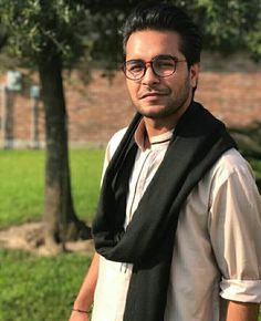 a man wearing glasses and a scarf standing in front of a tree with his hand on his hip