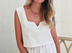 a woman with blonde hair wearing a white romper and gold necklace standing on a balcony