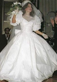 a woman in a white dress and veil walking down the aisle with her hand on her hip