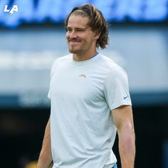 a man with long hair wearing a white t - shirt and blue shorts smiles at the camera