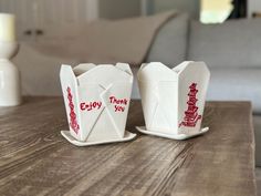 two white cups with red writing on them sitting on a wooden table next to a couch