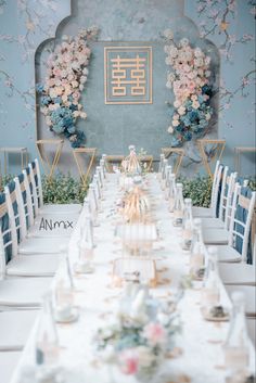 a long table with white chairs and floral decorations on the wall behind it is set for a formal dinner