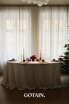 A Christmas-themed dining room with a festive table setting, a glowing Christmas tree, and sheer linen curtains in sand filtering soft natural light.