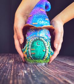 a person is holding a painted gnome's house on top of a wooden table