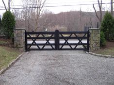 a driveway gate with stone pillars and an iron fence on the other side, in front of a wooded area