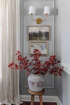 a vase with red flowers sitting on top of a stool in front of a window