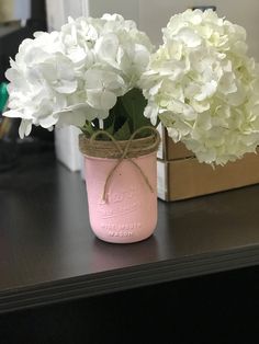 a pink mason jar filled with white flowers on top of a wooden table next to a brown box