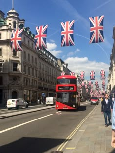 a red double decker bus driving down a street next to tall buildings with flags hanging from it's sides