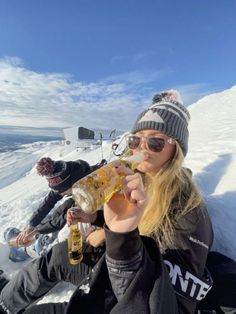 two people sitting in the snow and one is holding a bottle while another person takes a bite out of it